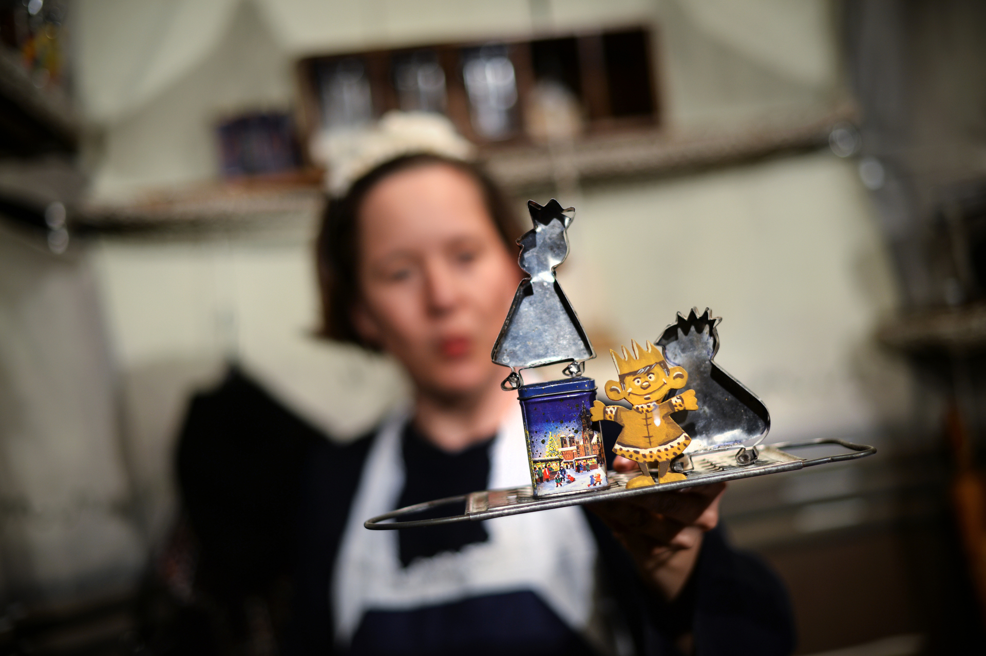 Frau mit weißer Schürze und weißem Häubchen hält ein Tablett mit Plätzchenförmchen vor sich