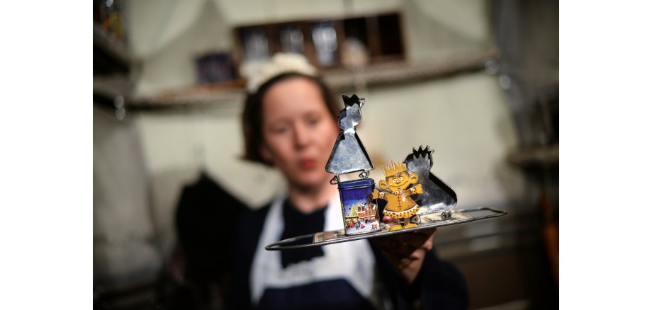 Frau mit weißer Schürze und weißem Häubchen hält ein Tablett mit Plätzchenförmchen vor sich